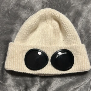 Vit ribbstickad mössa från C.P. Company - Vit ribbstickad mössa från C.P. Company med klassisk uppvikt kant och svarta glasögon-detaljer framtill. Tillverkad i 100% bomull för en mjuk och skön känsla. Perfekt accessoar för att lyfta din vinterstil.