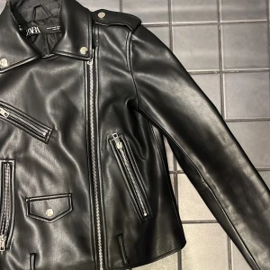 Svart skinnjacka från Zara - Snygg svart skinnjacka från Zara med flera silvriga dragkedjor och knappar. Jackan har klassisk bikerstil med axelklaffar och fickor med lock. Materialet är syntetiskt skinn och insidan är fodrad i svart tyg.