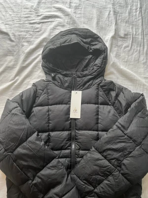 Svart pufferjacka från C.P. Company - Svart pufferjacka från C.P. Company med huva och dragkedja framtill. Jackan har quiltad design, två sidofickor med dragkedja och en ikonisk linsdetalj på ärmen. Perfekt för kalla dagar och har en clean, modern look.