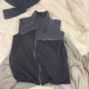 Svart Kingsland väst med dragkedja - Svart väst från Kingsland med blå detaljer och logga på ryggen. Övre delen är i glansig syntet och resten i mjuk fleece.