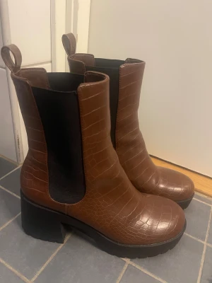 Bruna chelsea boots från Monki - Snygga bruna chelsea boots från Monki med croco-mönstrad yta och kraftig platt sula. Bootsens material är veganskt syntet och de har rund tå samt elastiska paneler på sidorna för enkel påtagning. Perfekt för dig som gillar chunky stil och detaljer.