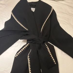 Svart kappa med broderad kant från Kappahl - Svart kappa från Kappahl med breda slag och snygga ljusa broderade detaljer längs kanterna. Jackan har ett knytbälte i midjan och lång ärm. Perfekt för dig som gillar stilrena och unika plagg med lite edge.