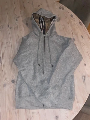 Burberry zip hoodie  - Snygg grå hoodie från Burberry med klassiskt rutigt foder i huvan. Tröjan har dragkedja framtill, snörning vid huvan och ribbade muddar. Perfekt för dig som gillar stilrena och bekväma plagg med en touch av lyx.