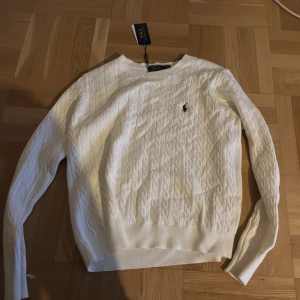 Beige kabelstickad tröja Polo Ralph Lauren - Vit kabelstickad tröja från Polo Ralph Lauren med klassisk rund hals och broderad logga på bröstet. Tröjan har långärmad passform och ribbade muddar vid hals, ärmslut och nederkant. Perfekt för dig som gillar stilrena och tidlösa plagg.