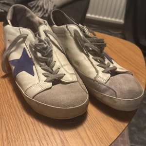 Golden Goose vita sneakers med blå stjärna - Golden Goose sneakers i vitt skinn med grå mockadetaljer på tån och grå snörning. Sidorna har en blå stjärna och hälen är svart med logotyp. Platt sula och rund tå, klassisk streetstil med coola detaljer.
