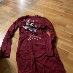 Vinröd Disneyklänning med Minnie Mouse - Vinröd långärmad klänning från Disney med tryck av Minnie Mouse och texten 'Waiting for Santa'. Klänningen har paljettrosett på motivet och är i mjuk bomull. Perfekt för julens mysiga dagar.