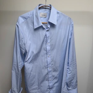 Blårandig skjorta från SANDO - Snygg skjorta från SANDO med tunna blå och vita ränder. Klassisk krage, långärmad och knäppning framtill med vita knappar. Skjortan har en stilren och fräsch look, perfekt för dig som gillar en clean och modern stil. Tillverkad i mjuk bomull för skön känsla hela dagen.