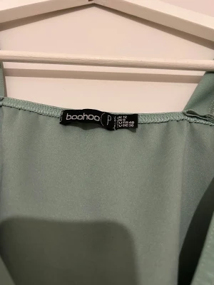 Grön omlottklänning från Boohoo - Snygg grön omlottklänning från Boohoo med korta ärmar och volangdetalj vid halsringningen. Klänningen har v-ringad front och ett knytband i midjan som ger en fin siluett. Perfekt för dig som gillar stilrena och trendiga plagg.