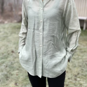 Ljusgrön oversized skjorta med knappar - Snygg ljusgrön skjorta i oversized modell med klassisk krage och knappar framtill. Skjortan har långa ärmar med manschett och är tillverkad i ett lätt, lite glansigt material som ger en chill vibe. Perfekt att styla med svarta jeans eller över en t-shirt.