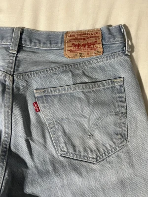 Levi's 501 ljusblå jeans straight fit - W34 L34, Ljusblå Levi's 501 jeans med klassisk straight fit och fem fickor. Jeansen har den ikoniska röda Levi's-taggen på bakfickan och patch i brunt läder i midjan. Tillverkade i slitstark bomullsdenim med knappgylf och normal passform. Säljer pga att jag inte använder dom