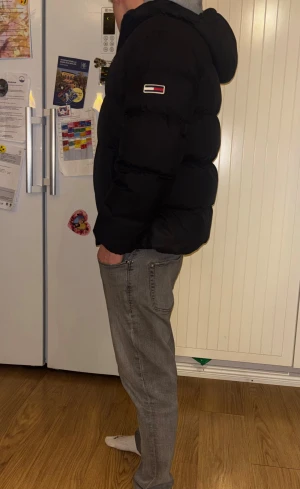 Svart pufferjacka Tommy Jeans M - Svart pufferjacka från Tommy Jeans med huva och broderade loggor på bröstet och ärmen. Jackan har dragkedja framtill med snygga detaljer i rött och vitt. Fylld för extra värme och har en loose passform, perfekt för kalla dagar.