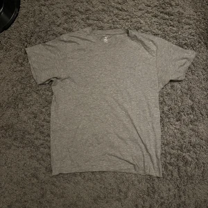 Grå t-shirt från SOC i bomull - En enkel och stilren grå t-shirt från SOC i storlek M. T-shirten har korta ärmar och är tillverkad i mjuk bomull, perfekt för träning eller chill. Den har en klassisk rund hals och en neutral grå färg som passar till allt.