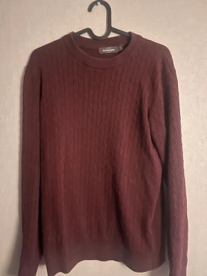 Vinröd stickad tröja Dressmann S - Snygg vinröd stickad tröja från Dressmann i storlek S. Tröjan har ett klassiskt kabelstickat mönster och rund halsringning. Perfekt för dig som gillar stilrena och tidlösa plagg med lite extra struktur. Passar dig som vill ha en enkel men ändå cool look.