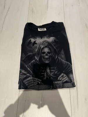 Svart t-shirt med dödskallemotiv - Säljer en svart t-shirt med stort tryck av en skelettfigur i kåpa som sitter vid ett bord med fjäderpenna, bok och ljusstake. Kedjor och dödskallar syns i bakgrunden. T-shirten har rund hals och korta ärmar. Perfekt för dig som gillar mörka och unika motiv.