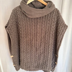 Brun stickad poncho  - Säljer en chunky brun stickad poncho från Cream Deluxe med stor krage och snygga träknappar på sidorna. Den har ett grovt flätmönster och är perfekt för att hålla sig varm under höst och vinter. Passar dig som gillar loose fit och mysiga vibes.