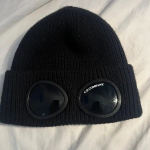 Svart C.P. Company mössa med goggles - Svart ribbstickad mössa från C.P. Company med ikoniska goggles framtill. Mössan är tillverkad i mjukt stickat material och har en uppvikt kant. Perfekt för dig som vill sticka ut med en streetwear-vibe och gillar coola detaljer.använd 2-3 gånger