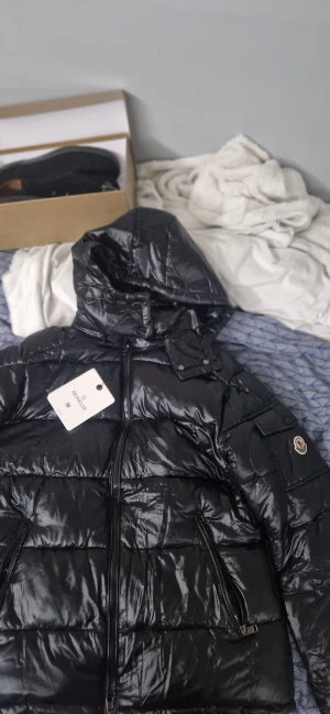 Svart pufferjacka från Moncler - (Helt ny och oanvänd)Säljer en svart pufferjacka från Moncler med klassisk logotyp på bröstet och coolt serietryck på insidan. Jackan har blanka ytor, blå och röda detaljer och knappar med Moncler-gravyr. Perfekt för dig som vill ha en snygg och varm jacka med exklusiv känsla.
