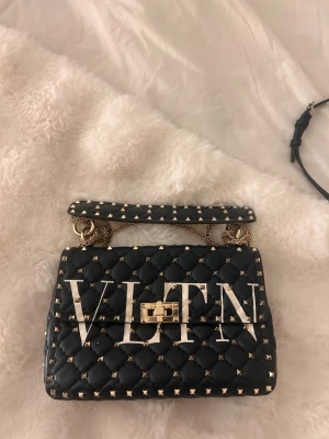 Svart quiltad handväska från Valentino - Snygg svart handväska från Valentino med stora vita VLTN-bokstäver på framsidan. Väskan är quiltad och dekorerad med guldfärgade nitar och har ett guldfärgat lås. Kedjehandtag och exklusiv design som verkligen sticker ut. INGEN BESTÄLLNINGSVARA!