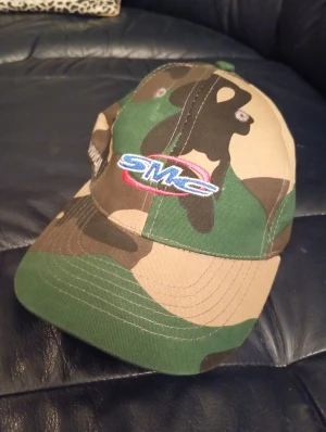 SMC keps med camouflage-mönster - Snygg keps från SMC med klassiskt camouflage-mönster i grönt, beige och brunt. Framtill finns en broderad SMC-logga i blått, vitt och rött. Perfekt för dig som vill sticka ut med streetstyle eller gillar outdoor-vibbar.