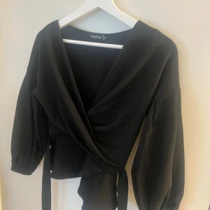 Svart omlottopp med knyt från Boohoo - Svart omlottopp från Boohoo med v-ringad front och breda, puffiga långärmar. Toppen har ett snyggt knyt i midjan som ger en markerad siluett och volangkant nedtill. Materialet är mjukt och har en lätt struktur, perfekt för att lyfta din outfit.