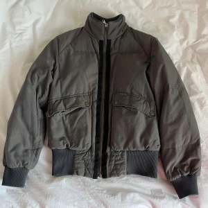 Grå bomberjacka från Dolce & Gabbana - Snygg grå bomberjacka från Dolce & Gabbana med ribbade muddar, hög krage och dragkedja framtill. Jackan har två fickor med lock och knappar, samt coola patchdetaljer på ärmen och insidan. Perfekt för dig som gillar streetstyle och exklusiva märken🩶