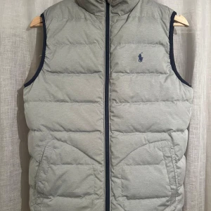 Grå och marinblå dunväst från Polo Ralph Lauren - Två snygga dunvästar från Polo Ralph Lauren, en i ljusgrått och en i marinblått. Båda har quiltad design, dragkedja framtill och broderad logga på bröstet. Perfekt för lager-på-lager och chill stil. Fickor med dragkedja och hög krage för extra värme. Den är i storlek L (14-16) och har ett litet hål.