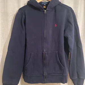Mörkblå hoodie från Polo Ralph Lauren - Mörkblå hoodie från Polo Ralph Lauren med dragkedja framtill och klassisk röd broderad logga på bröstet. Tröjan har huva, två fickor och långa ärmar. Perfekt för chill dagar och enkel att matcha med jeans eller joggers.