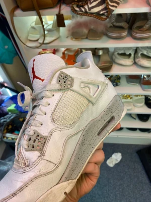 Nike Air Jordan 4 vita sneakers - Nike Air Jordan 4 sneakers i vitt skinn med grå detaljer och stänkmönstrad sula. Klassisk meshpanel på sidorna, snörning och röd Jumpman-logga på plösen. Snygg retrostil med platt sula och rund tå, perfekt för dig som gillar streetwear.