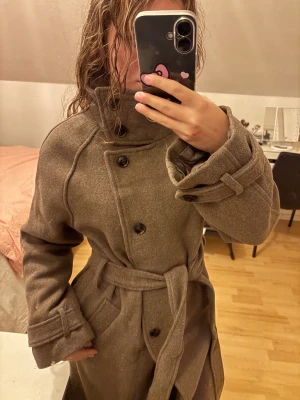 Belted wool blend coat GINATRICOT  - Säljer en lång brun kappa med bred krage och bälte i midjan. Kappan har knappar framtill, långa ärmar med snygga detaljer vid handlederna och en rak, klassisk passform. Perfekt för dig som vill ha en stilren och tidlös look . Köpte denna pch en svart dock i xs då det var de enda kvar men ingen passar, de är slutsålda helatiden. o returtiden är över pga utlandspraktik jag hade. Två reservknappar tillföljer.128cm lång Helt ny! Prislapp kvar