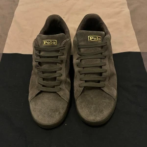 Grå mocka sneakers från Polo Ralph Lauren - Snygga sneakers i grå mocka från Polo Ralph Lauren med diskret logga på sidan och guldiga Polo-detaljer på plös och häl. Skorna har rund tå, platt sula och klassisk snörning. Perfekta för dig som gillar stilrena och tidlösa sneakers med premiumkänsla.