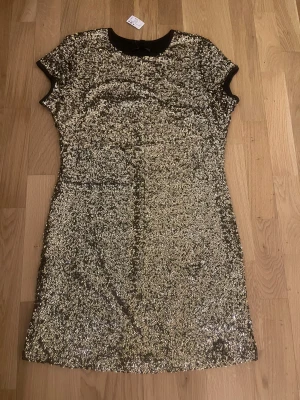 Guldig paljettklänning från Mango - Supersnygg kort klänning från Mango, täckt med glittrande guldiga paljetter. Modellen är figurnära med rund hals och korta ärmar, perfekt för att sticka ut på festen. Materialet är polyester och klänningen har en lyxig känsla med sin glittriga yta.