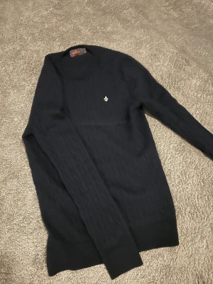 Svart kabelstickad tröja Morris M - Svart kabelstickad tröja från Morris i 100% merinoull. Rund hals och diskret Morris-logga broderad på bröstet. Långärmad och klassisk passform, perfekt för dig som gillar stilrena och tidlösa plagg. Den är sitter som xs inte M