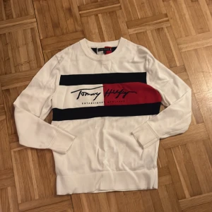 Vit Tommy Hilfiger tröja med logga - Vit långärmad tröja från Tommy Hilfiger i storlek S. Framsidan har ett stort tryck med logga i svart, rött och marinblått. Tröjan har rund hals och ribbade muddar vid ärmslut och nederkant. Perfekt för dig som gillar klassisk streetstyle.