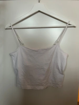 Vit linnetopp med smala axelband - Säljer en enkel och stilren vit linnetopp med smala justerbara axelband. Toppen har en rak och croppad passform som är perfekt för varma dagar. Materialet är mjukt och lätt bomull som känns skönt mot huden. Perfekt att styla med jeans eller kjol.