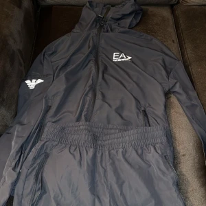 EA7 mörkblå träningsdräkt, storlek S - En snygg träningsdräkt från EA7 Emporio Armani. Dräkten är i en mörkblå färg och har en sportig design med logotypen på bröstet och armen.   Den är knappt använd och är i nyskick!