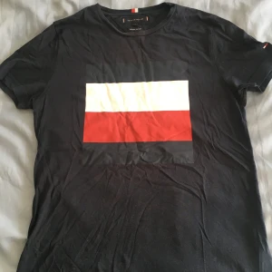 Mörkblå t-shirt från Tommy Hilfiger - Snygg mörkblå t-shirt från Tommy Hilfiger i ekologisk bomull. Den har en stor vit och röd rektangel på framsidan som ger en cool look. Passar bra till jeans!