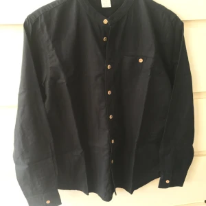 Mörkblå skjorta Kids från Lindex - Säljer en stilren mörkblå skjorta från Lindex i storlek 164. Skjortan har långa ärmar och knappar i träfärg som ger en snygg kontrast. Perfekt för både vardag och festliga tillfällen. Passar bra till jeans eller chinos för en avslappnad look.