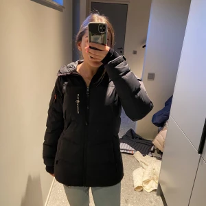 Svart dunjacka från Sail Racing - Säljer en svart dunjacka från Sail Racing i mycket bra skick. Jackan har en dragkedja framtill och är långärmad med en skön huva. Perfekt för kalla vinterdagar och höstpromenader. Den är både stilren och funktionell, vilket gör den till ett måste i garderoben!