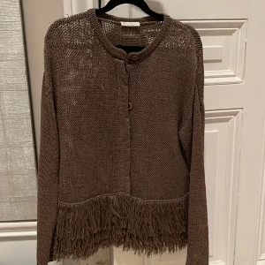 Brun stickad kofta från Brunello Cucinelli - Säljer en superfin brun stickad kofta från Brunello Cucinelli. Den har långa ärmar och en unik fransdetalj längst ner som ger den en bohemisk elegant touch. 