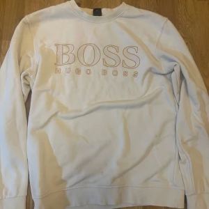 Vit sweatshirt från Hugo Boss - Säljer en stilren vit sweatshirt från Hugo Boss med deras ikoniska logga tryckt på bröstet. Tröjan har långa ärmar och en klassisk rund halsringning. Perfekt för en avslappnad och trendig look. Passar mellan M-L