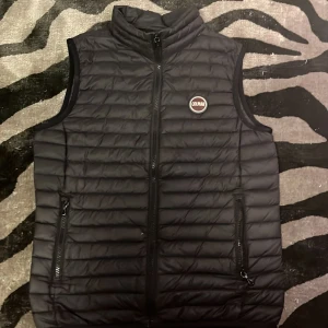 Svart dunväst från Colmar - Säljer en stilren svart dunväst från Colmar med dragkedja och hög krage. Västen har två praktiska fickor med dragkedjor och en diskret logga på bröstet. Perfekt för lager på lager och även på våren,     Nypris:2500