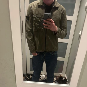 Stone Island overshirt - Säljer en snygg grön jacka från Stone Island med dragkedja och en ficka på bröstet. Jackan har en klassisk look med långärmade ärmar och en ikonisk logga på ärmen. Perfekt för en stilren och avslappnad stil. Nypris runt 2500kr bara höra av sig vid eventuella frågor!