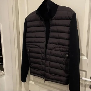 Svart quiltad jacka från Moncler - Snygg svart quiltad jacka från Moncler med dragkedja och långa ärmar. Jackan har en stilren design med ribbad krage och två praktiska fickor med dragkedja framtill. Perfekt för kyligare dagar.