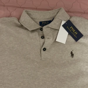 Grå långärmad pikétröja från Polo Ralph Lauren - Säljer en stilren grå långärmad pikétröja från Polo Ralph Lauren. Tröjan har en klassisk krage med knappar och det ikoniska broderade logotypen på bröstet. Perfekt för en avslappnad men ändå elegant look.