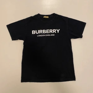 Svart Burberry London T-shirt - Säljer en snygg svart t-shirt från Burberry med vit text 'BURBERRY LONDON ENGLAND' på framsidan. Den är i bra skick och perfekt för en casual look. Passar både till vardags och fest. Den har två små fläckar på  r och e men inget som märks av vid användning, skriv vid minsta fråga eller fundering💯👍