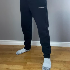 Svarta mjukisbyxor från Softwear UF - Säljer ett par svarta mjukisbyxor från Softwear UF. Byxorna har en avslappnad passform och är perfekta för en bekväm dag hemma. De har en elastisk midja och en enkel design med textdetalj på benet.