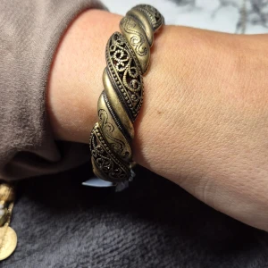 Antikguld armband med snirkligt mönster - Snyggt armband i antikguld med ett detaljerat snirkligt mönster. Armbandet har en vriden design som ger det en unik och elegant look. Perfekt för att ge en vintage touch till din outfit.