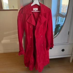 Röd trenchcoat - Snygg röd trenchcoat med knappar och bälte i midjan. Perfekt för att ge en färgklick till din outfit. Klassisk design med krage och långa ärmar.