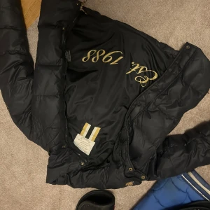 Svart dunjacka med broderad detalj - Snygg svart dunjacka med broderad detalj på bröstet och text på insidan. Jackan har en hög krage och stängs med dragkedja och knappar. Perfekt för kyligare dagar.