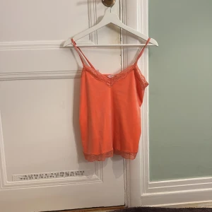 Orange linnetopp från Hunkemöller - Säljer en snygg orange linnetopp från Hunkemöller med spetsdetaljer vid halsringningen och nederkanten. Toppen har smala axelband och är perfekt för varma dagar.